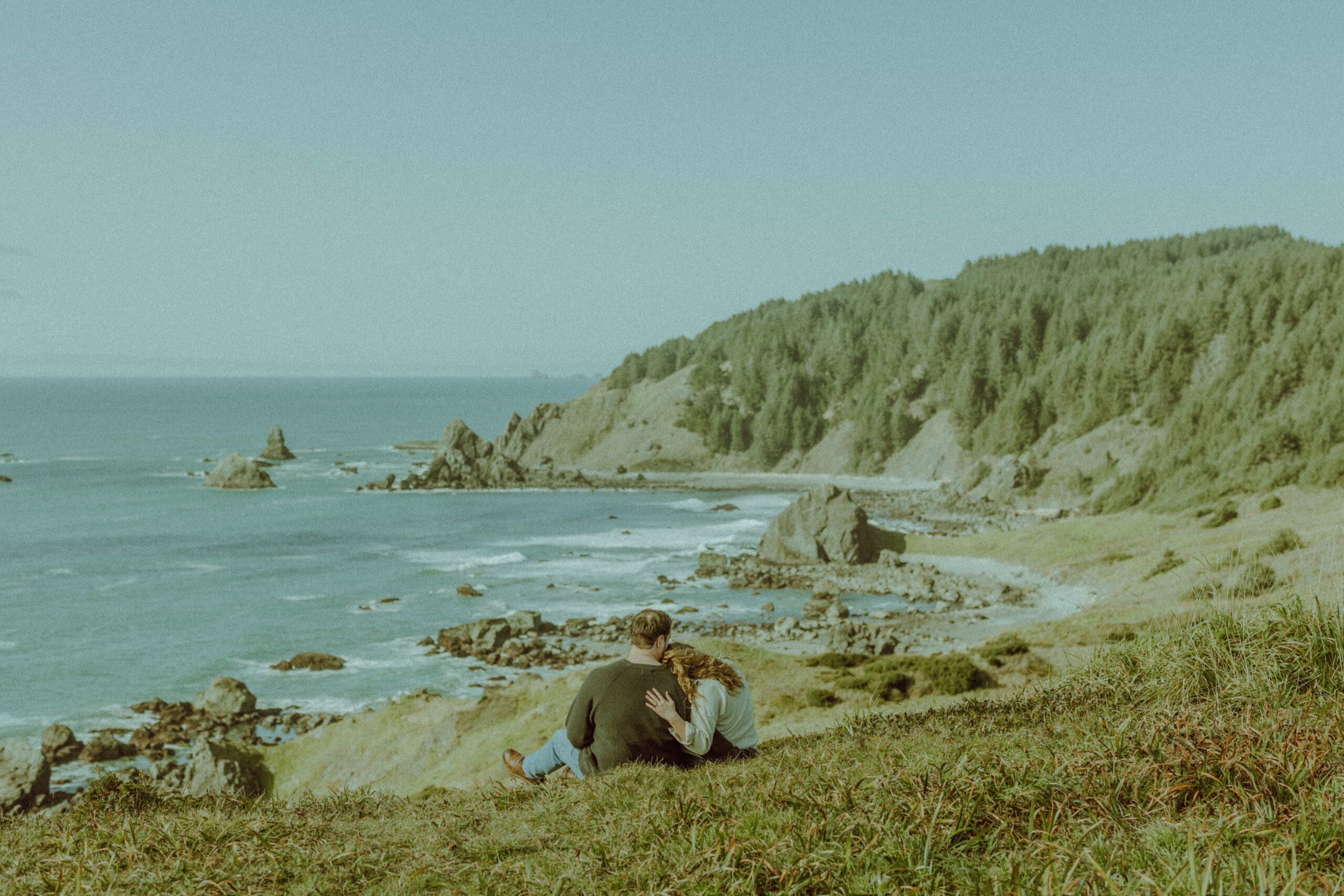 couple sitting on a grassy hill for their engagement photos at the oregon coast