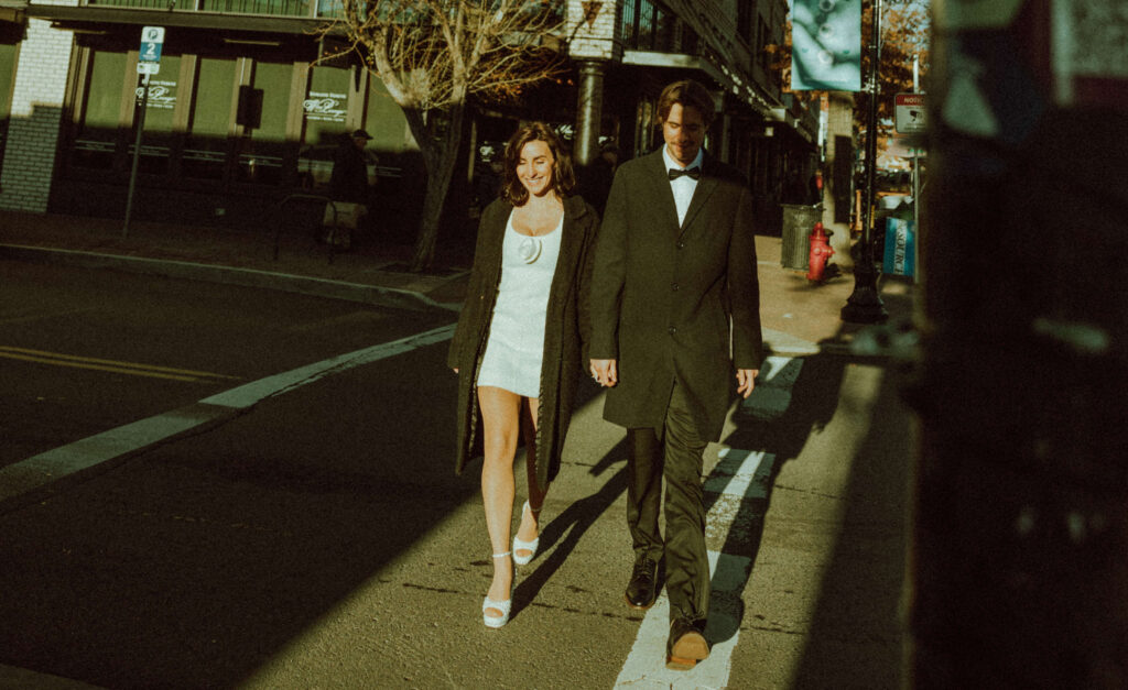 bride and groom holding hands walking across the cross walk in downtown bend oregon for their editorial engagement photos 