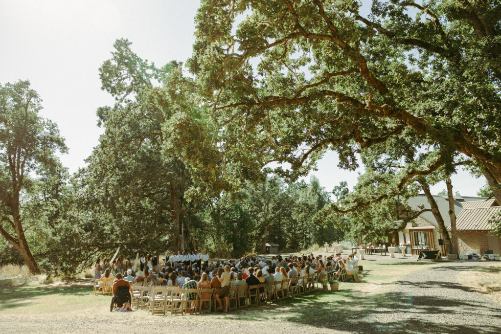 Outdoor wedding ceremony at Mt Pisgah Arboretum in Oregon, featuring a large gathering under sprawling trees.